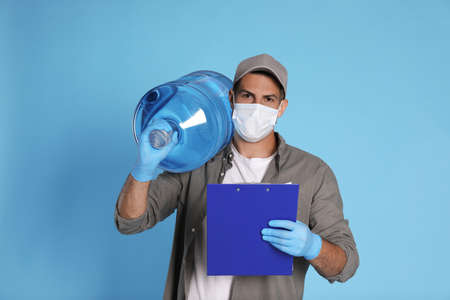 Courier in medical mask with bottle for water cooler and clipboard on light blue background. Delivery during coronavirus quarantineの写真素材