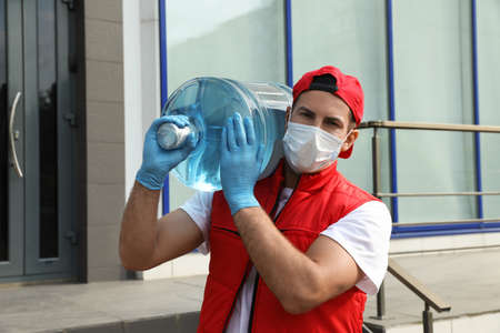 Courier in medical mask holding bottle for water cooler near building outdoors. Delivery during coronavirus quarantineの写真素材