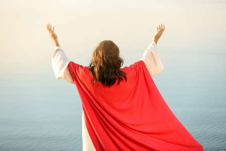 Jesus Christ raising hands near water outdoors, back viewの写真素材