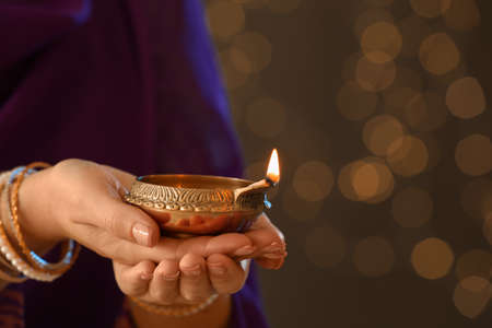Woman holding lit diya lamp in hands against blurred lights, closeup. Diwali celebrationの写真素材