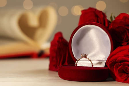 Beautiful engagement ring with gemstone in box and flowers on white wooden table. Space for text