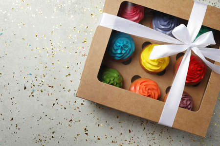 Box with delicious colorful cupcakes and confetti on light gray table, flat lay. Space for text
