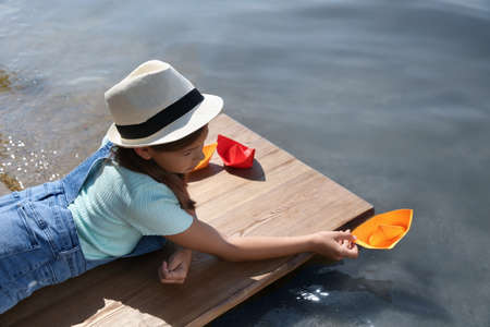 Cute little girl playing with paper boat on wooden pier near riverの写真素材