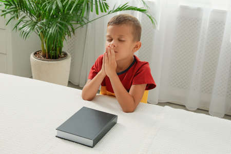 Cute little boy with hands clasped together saying prayer over Bible at homeの写真素材