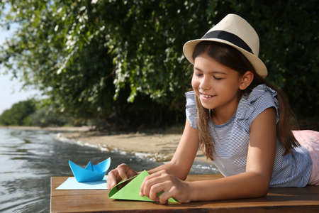 Cute little girl making paper boats on wooden pier near riverの写真素材