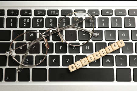 Wooden cubes with word Blacklist and glasses on laptop keyboard, top view