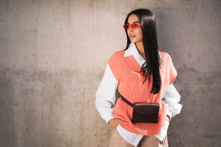 Beautiful young woman with stylish waist bag near gray wall outdoors, space for text