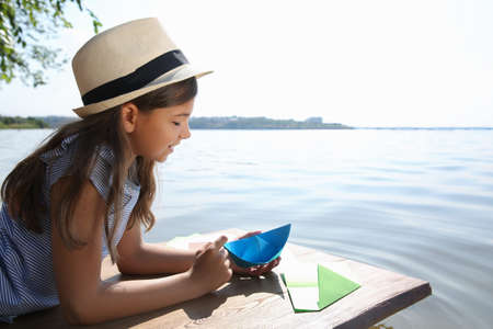 Cute little girl making paper boats on wooden pier near riverの写真素材