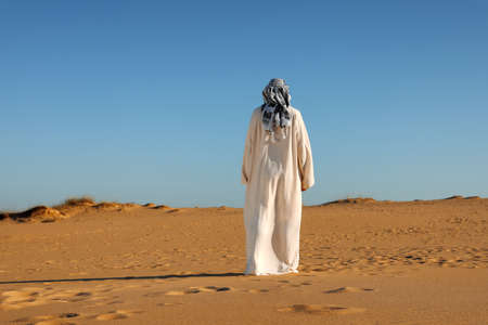 Man in arabic clothes walking through desert on sunny day, back viewの写真素材