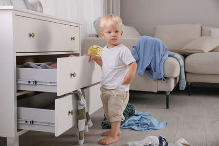 Cute little boy playing with clothes in dresser's drawer at home