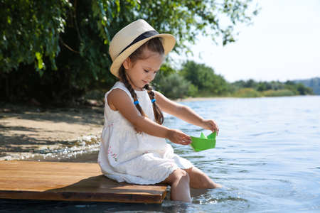 Cute little girl playing with paper boat on wooden pier near riverの写真素材