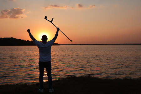 Man raising elbow crutch up to sky near river at sunset, back view. Healing miracleの写真素材