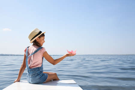 Cute little girl playing with paper boat on wooden pier near riverの写真素材