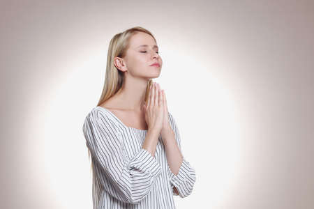 Religious young woman with clasped hands praying against light background. Space for text