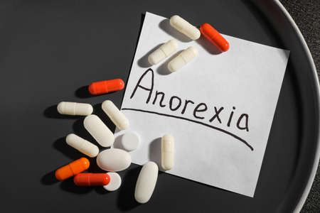 Pills and note paper with word Anorexia on gray plate, flat lay
