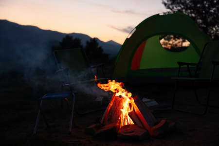Beautiful bonfire and folding chairs near camping tent outdoors in eveningの写真素材