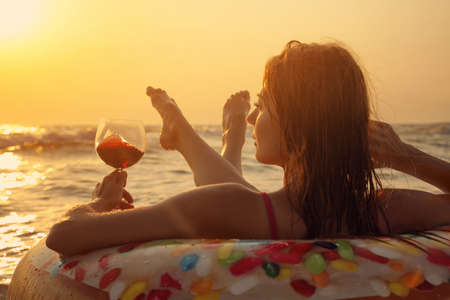 Woman with glass of wine and inflatable ring in sea at sunsetの写真素材
