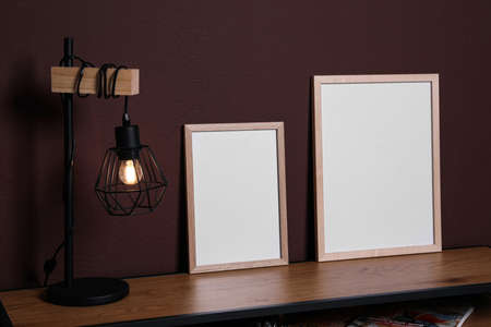 Empty frames and stylish lamp on wooden table near brown wall. Mockup for designの写真素材