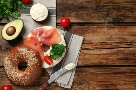 Delicious bagel with cream cheese, jamon, tomato and parsley on wooden table, flat lay. Space for textの写真素材