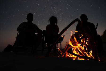 Group of friends gathering around bonfire in evening, focus on flame. Camping season