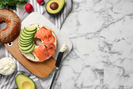 Delicious bagel with cream cheese, salmon and avocado on white marble table, flat lay. Space for...