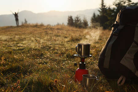 Camping burner with mug of hot drink near backpack on grass in mountains. Space for textの写真素材