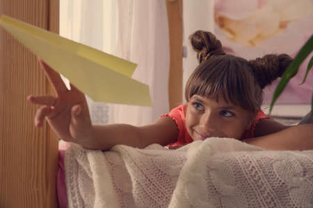 Cute little girl playing with paper plane on bed in roomの写真素材