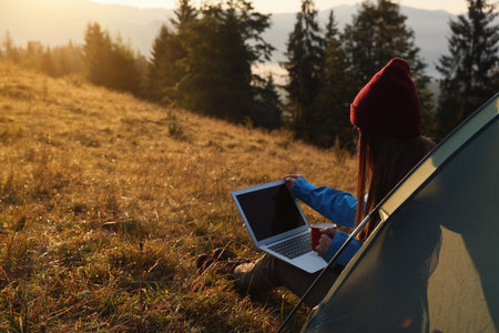 Woman working on laptop near camping tent outdoors surrounded by beautiful natureの写真素材