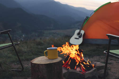 Yellow mug with hot drink on wooden stump near bonfire outdoors. Camping seasonの写真素材
