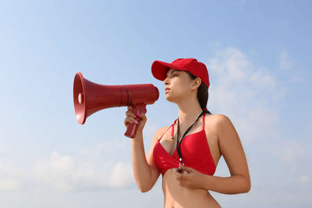 Beautiful young lifeguard with megaphone against blue skyの写真素材