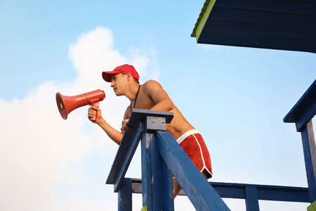 Handsome lifeguard with megaphone on watch tower against skyの写真素材