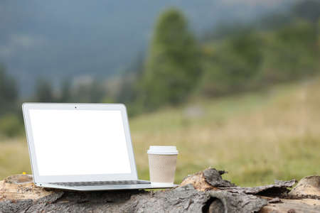 Modern laptop with blank screen and coffee cup on tree trunk in nature, space for text. Working outdoorsの写真素材