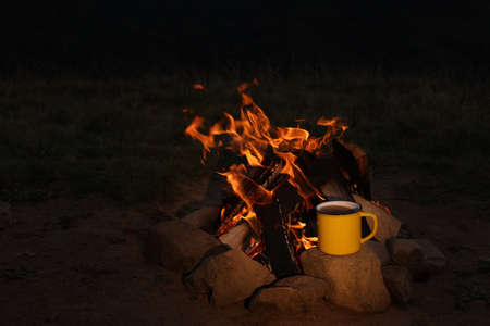 Metal mug with drink near beautiful bonfire outdoors at night. Camping seasonの写真素材