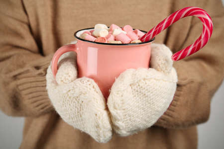 Woman in knitted mittens holding cup of delicious hot chocolate with marshmallows and candy cane, closeupの写真素材