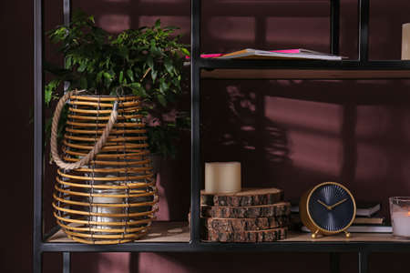 Stylish holder with burning candle, houseplant and decor on shelving unit near brown wallの写真素材