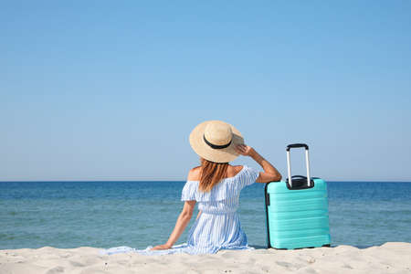 Woman with suitcase sitting on sandy beach near sea, back viewの写真素材