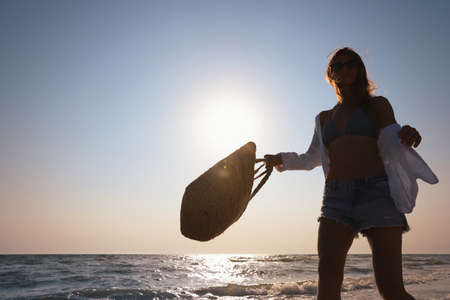 Woman with beach bag walking on sunlit seashoreの写真素材