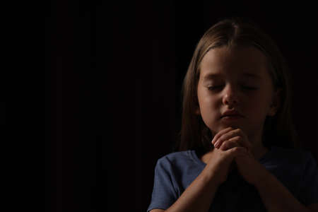 Cute little girl with hands clasped together praying on black background. Space for textの写真素材