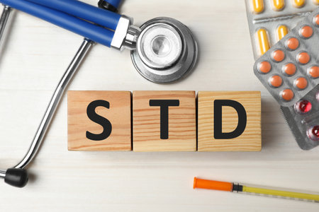 Abbreviation STD made with cubes, drugs and stethoscope on white wooden table, flat lay