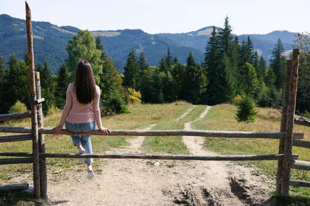 Young woman sitting on wooden fence and enjoying mountain landscape, back view. Space for textの写真素材