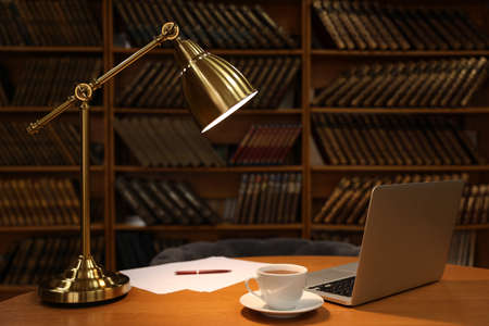Lamp, cup of drink and laptop on wooden table near shelves with collection of vintage books in libraryの写真素材