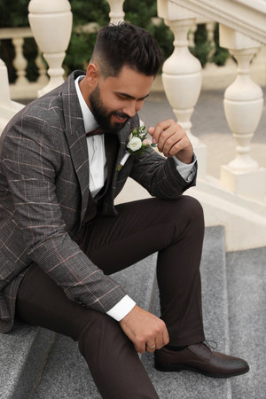 Young groom in elegant suit with beautiful boutonniere sitting on stairs outdoorsの写真素材