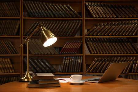Lamp, cup of drink and laptop on wooden table near shelves with collection of vintage books in libraryの写真素材