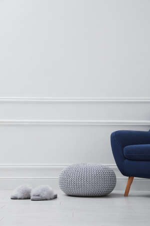 Knitted pouf, fuzzy slippers and armchair near white wall indoors. Space for textの写真素材