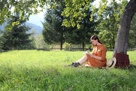 Beautiful young woman drawing with pencil in notepad near tree on green grass