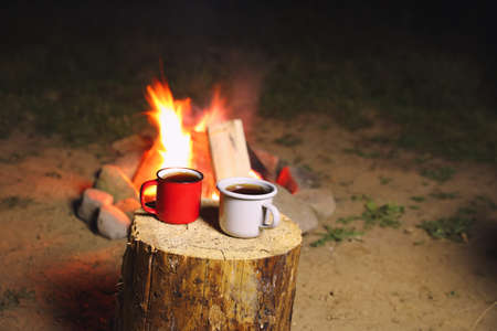 Cups of hot drink on old log near bonfire outdoors. Camping seasonの写真素材
