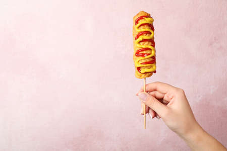Woman holding delicious corn dog with mustard and ketchup on pink background, closeup. Space for...