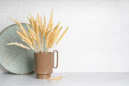 Ceramic vase with fluffy dry plants and decorative plate on light table near white brick wall. Space for textの写真素材