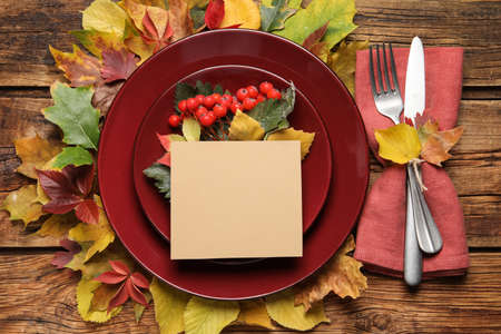 Festive table setting with autumn decor and blank card on wooden background, flat lay