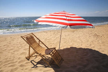 Deck chair near red and white striped beach umbrella on sandy seashoreの写真素材